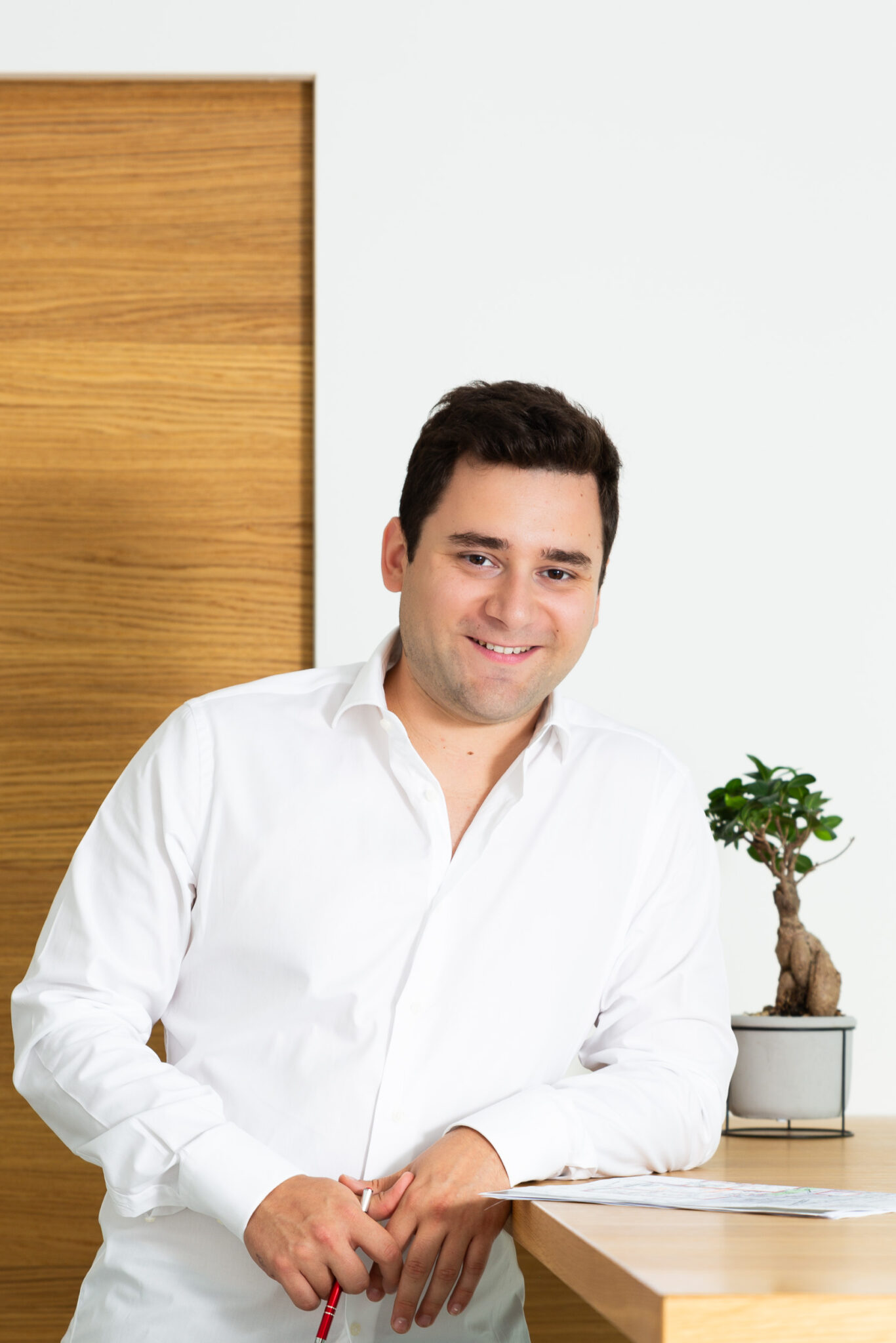 Junger Mann in weißem Hemd steht an einem Schreibtisch mit einer kleinen Pflanze, lächelt freundlich in die Kamera.