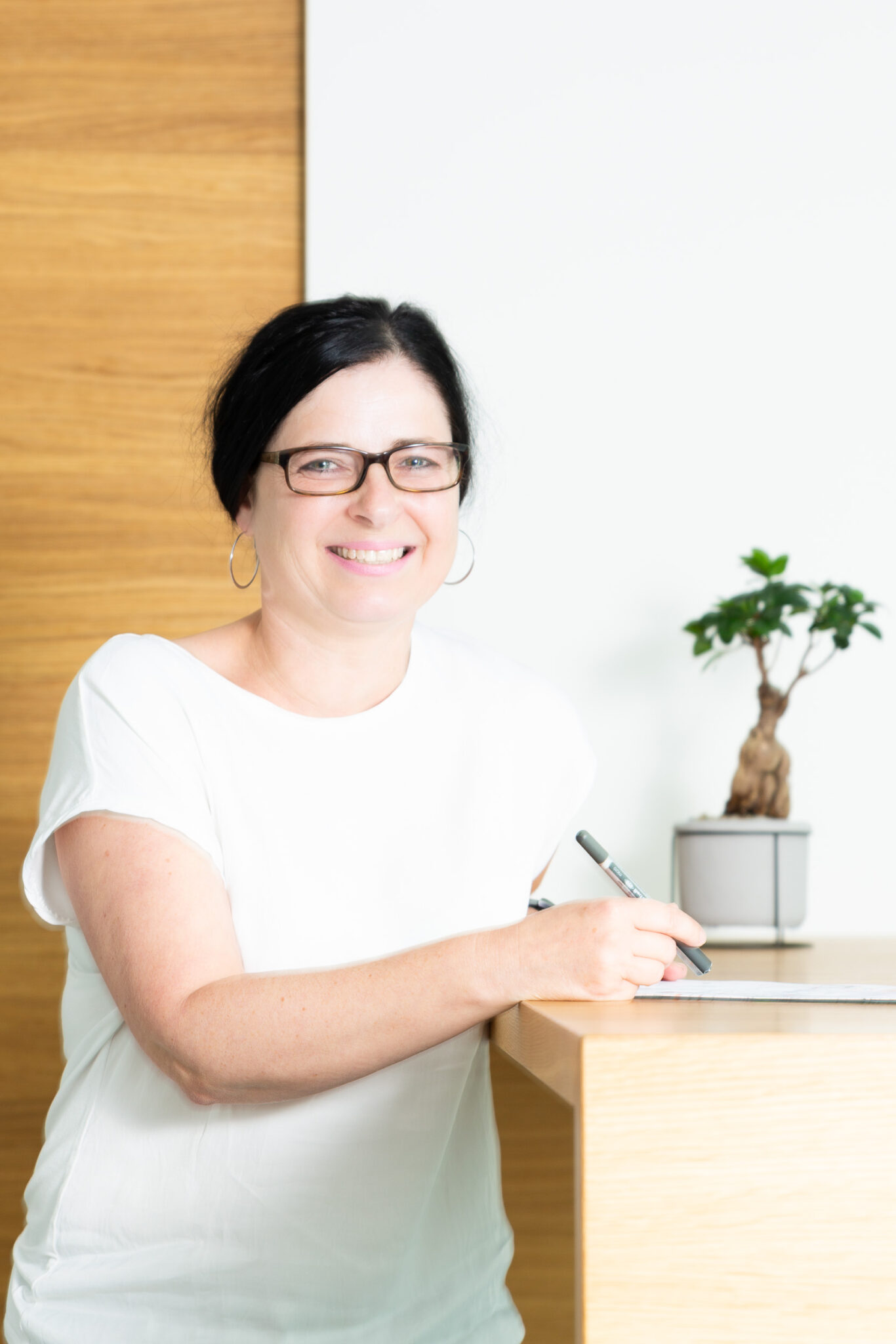 Frau mit Brille und schwarzem Haar, die an einem Schreibtisch sitzt und einen Stift hält, neben einem kleinen Bonsai-Baum.