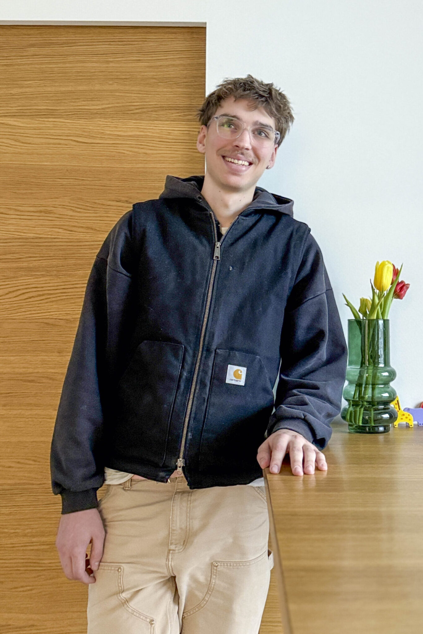 Junger Mann mit Brille, der an einem Holztisch steht und eine schwarze Jacke trägt, mit einer Vase voller Blumen im Hintergrund.