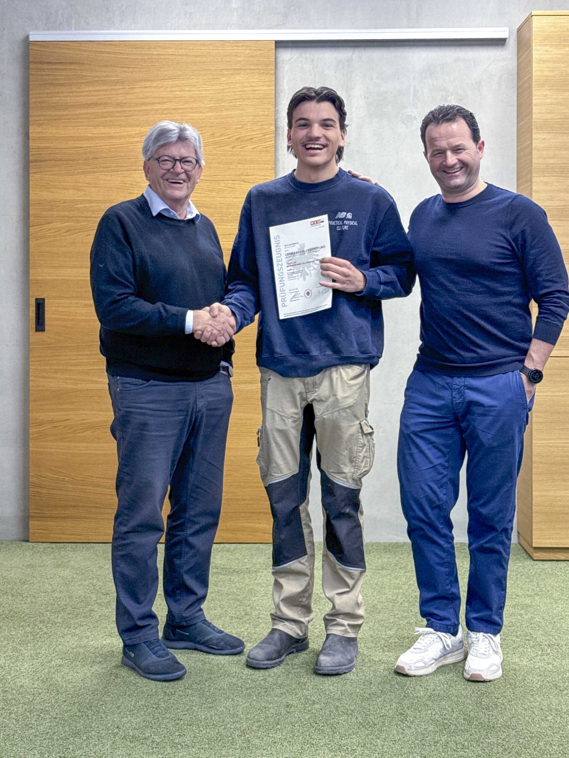 Drei Personen stehen zusammen und halten ein Zertifikat in einer modernen Innenumgebung.